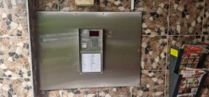Brushed steel intercom panel with numeric keypad and small red display mounted on a mosaic-tiled wall; magazine rack to the right.